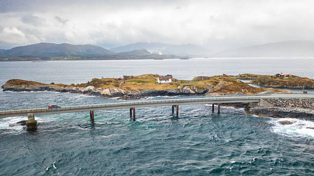Dramatic engineering of Atlantic ocean road weaves across small rugged islands. Road stretches among calm waters to foggy distance under overcast sky