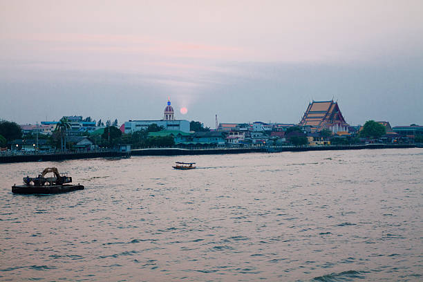 Sunset and sun over Chao phraya river in Bangkok.  View from eastern riverside towards north with cityscape along the riversides. On opposite side are a thai temple and  a church with illuminated cross on cupola. Church is Santa Cruz Church in Khwaeng (sub-district) Wat Kanlaya of the Thon Buri District