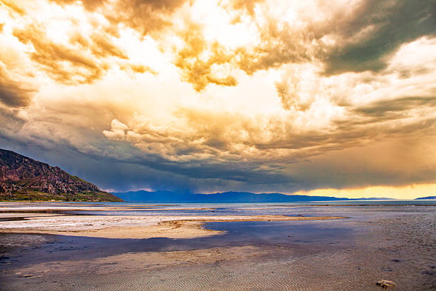 A dramatic golden sky dominates this panoramic shot of a salt flat, with storm clouds rolling over distant mountain ranges. The rippled patterns of the salt crust and subtle pools of water reflect the moody atmosphere of an impending storm. The golden hue of the clouds suggests either early sunrise or late evening light, adding cinematic warmth to the threatening sky. Taken from the shoreline looking out over the flats, this photo captures the raw elemental beauty of weather over desolate terrain.