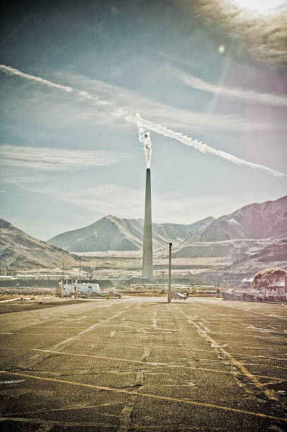 A lone industrial smokestack releases a thin plume against layered mountains and streaky high clouds. The foreground is an empty, weathered parking lot leading the eye toward the stack and the distant plant. Midday haze and light flare give the scene a muted, vintage feel, contrasting heavy industry with a vast western landscape.