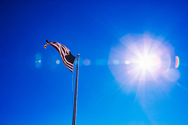 A U.S. flag waves from a silver pole against a vivid, cloudless blue sky. The sun sits at frame right, producing a strong starburst and a string of circular lens flares across the sky. Large negative space emphasizes the flag’s silhouette and motion. Daytime, clear weather. Static shot.