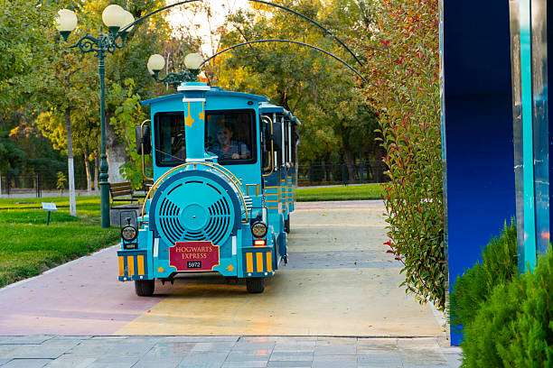 Tashkent, Uzbekistan, October 18, 2025: Bright turquoise amusement park train with yellow accents driving along a tree-lined path with autumn foliage and vintage street lamps