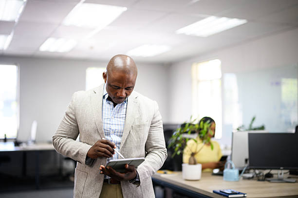 Business man looking down and reading information on a digital tablet in a brightly lit office, illustrating focus, responsibility and productivity at work.