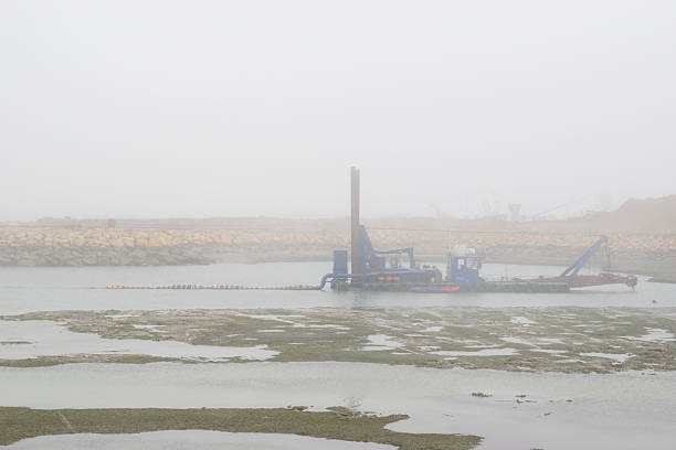 Industrial dredging barge working in a foggy harbor, with long suction pipeline floating on calm water and rocky shore in foreground, concept of marine construction, coastal engineering and port maintenance.
