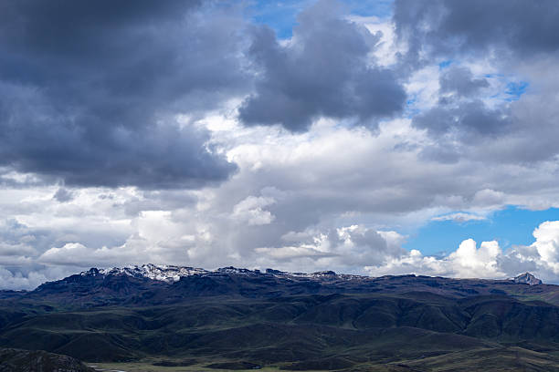 A rugged mountain range in Peru, partially covered with snow, sits beneath a dramatic sky filled with dark storm clouds and patches of blue