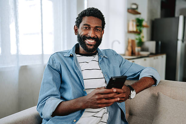 istock Smiling man sitting on sofa using smartphone at home looking at the camera 2243821104