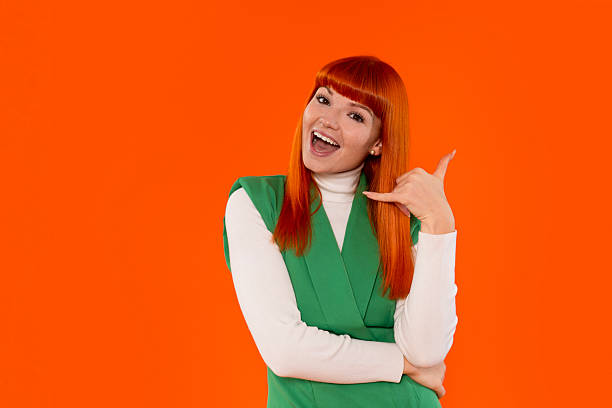 istock Cheerful woman making a phone gesture with a bright orange background in a studio setting 2243827559