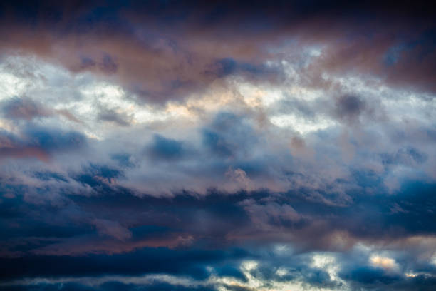 An abstract, close-up photograph of a dramatic, turbulent sky, with clouds colored in deep blues, purples, and warm pinks and whites from sunset or sunrise light.