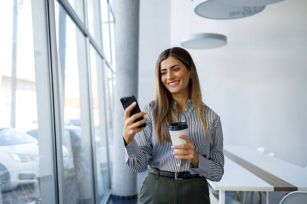 istock Businesswoman standing in modern office holding coffee and checking smartphone, smiling, using mobile app 2243831771