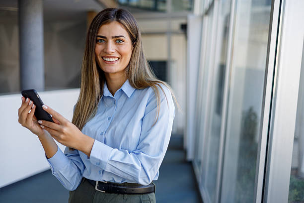 istock Professional woman smiling, holding smartphone in modern office building corridor, using technology for business 2243833618