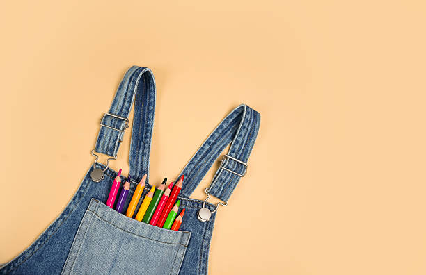 Brightly colored pencils are arranged neatly in the pocket of denim overalls. The scene is set against a soft, neutral background, adding a creative touch to the image.