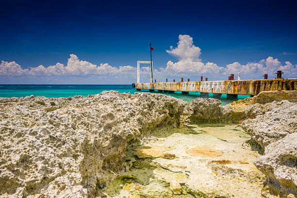 A horizontal photograph of a rugged, rocky shore in the foreground, with an old concrete pier extending into the turquoise ocean under a blue sky.