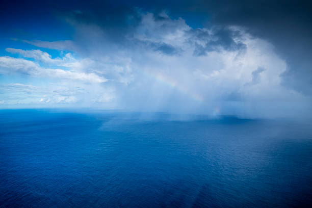 An aerial photograph of the vast, deep blue ocean. A rain shower in the distance creates a faint rainbow under dramatic, dark, and white storm clouds.