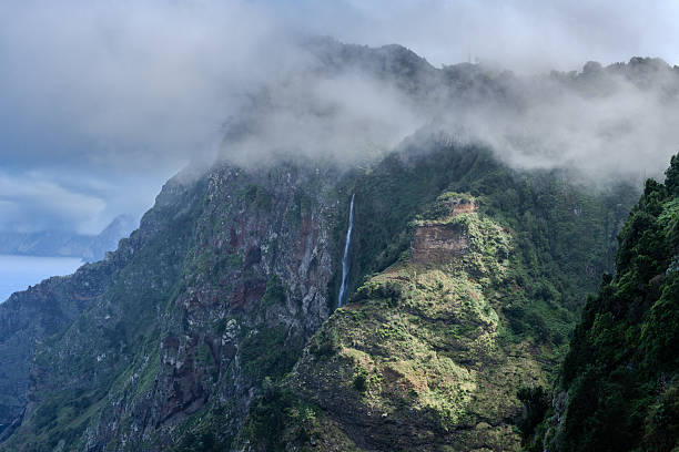 A majestic waterfall cascades through the lush green mountains of Madeira, surrounded by dense vegetation. Misty clouds hover over the peaks, creating a dramatic and serene alpine atmosphere on the island.