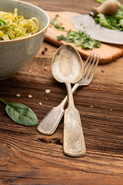 Old cutlery placed on wooden table next to bowl of spaghetti with green pesto and fresh parsley.