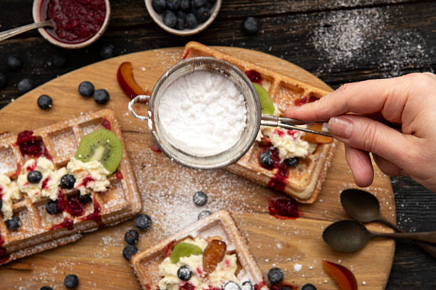 Close up of hand using sifter to sprinkle sugar over waffles topped with cream, blueberries and kiwi on rustic wooden board.