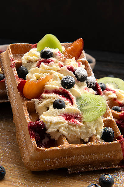 Crispy waffles topped with whipped cream, blueberries and kiwi slices, decorated with sugar on rustic background.