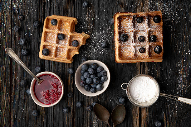 Homemade Belgian waffles sprinkled with powdered sugar, topped with fresh blueberries and served with raspberry jam on rustic dark wooden background.