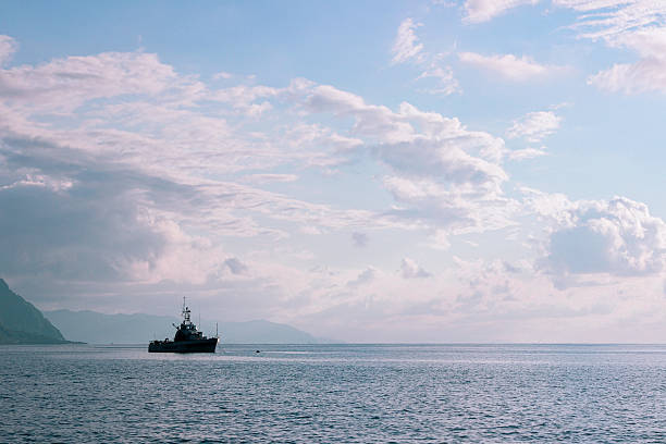 A lonely ship rests on the water against a calm sea horizon. Distant mountains are visible while the sky is decorated with soft clouds, creating an atmosphere of coziness and serenity