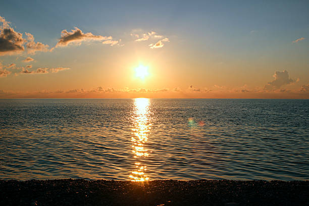 The warm glow of the sunset floods the horizon and reflects on the calm sea surface, creating a sense of peace and harmony. Clouds gently frame the sunlight