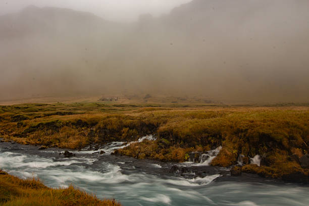 ​A long exposure shot of the Fossalar River cascading over dark rocks, creating a silky smooth water effect. In the distance, a small, isolated structure sits within a vast, golden-brown landscape shrouded in thick fog and low clouds, characteristic of Iceland's wild and dramatic natural beauty.