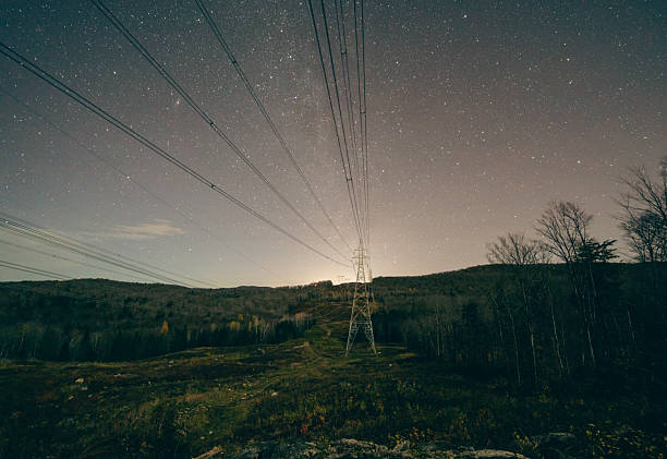 A tranquil night scene shows a star-filled sky above rolling hills, with a lattice of power lines and a lone transmission tower cutting across the quiet landscape.