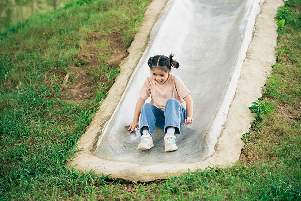 A cheerful girl slides down a blue playground slide on a sunny autumn day, surrounded by trees and fallen leaves in a quiet park.