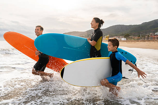 Family running through ocean waves carrying surfboards, wearing wetsuits, experiencing joy while engaging in water sports activity on a sunny beach day