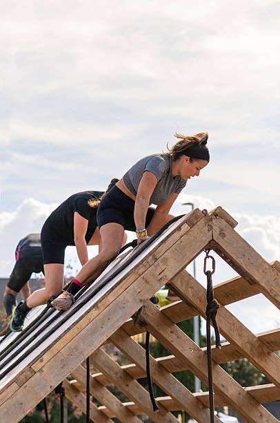 A determined female athlete and other competitors overcoming a wooden A-frame obstacle at its peak during an outdoor obstacle race.