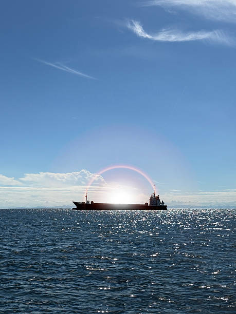 Cargo ship silhouetted against sunset with dramatic lens flare over sparkling ocean surfaceTags