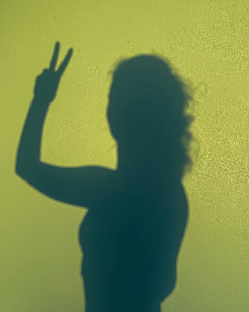 Silhouette of a girl with her hand raised with a peace symbol casting a shadow on a green wall in the sunset light.