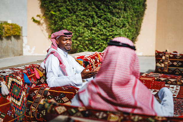 Middle Eastern men in traditional dress engaging in conversation at an outdoor Arabic Majlis setting. One man gestures while talking, seated on colorful patterned cushions, with a calm and friendly expression. The setting features outdoor seating and greenery, creating a relaxed atmosphere.