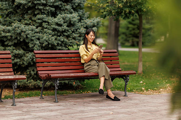istock Smiling Asian woman using smartphone while relaxing on a park bench 2243917966