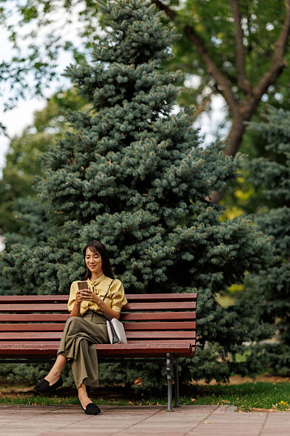 istock Smiling Asian woman using smartphone while relaxing on a park bench 2243919765