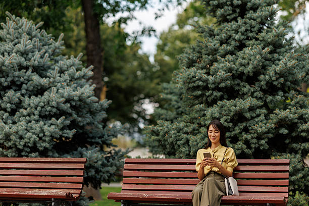 istock Smiling Asian woman using smartphone while relaxing on a park bench 2243919819
