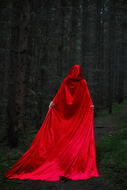 istock figure wearing a flowing red cloak stands on a narrow path in a dense forest. Tall trees surround the scene, and the light is dim, creating a mysterious atmosphere 2243922629