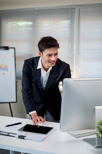 istock Asian businessman leaning over working table holding mouse and looking at computer in company office 2243924710
