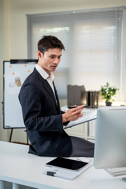 istock Asian businessman holding clipboard and looking at computer while sitting on table in company office 2243924753