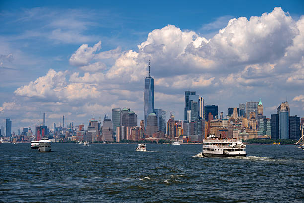 Scenic view of boats and a ferry crossing the Hudson River with the Manhattan skyline in the background. The One World Trade Center tower rises prominently among the skyscrapers under a dramatic summer sky filled with clouds.