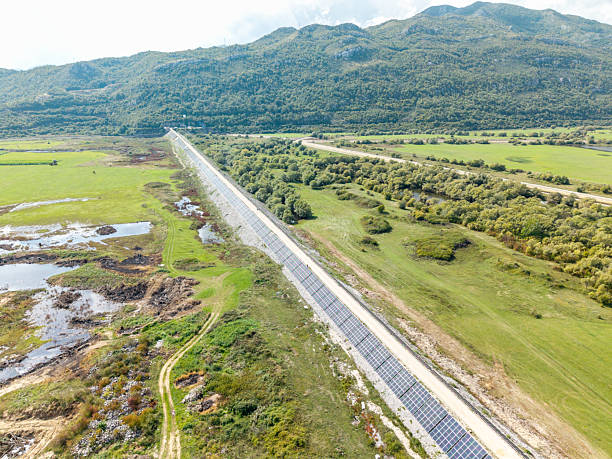 Long row of solar panels installed beside a dirt road in a green rural valley. Renewable energy technology in natural landscape with mountains in the background. Sustainable power generation concept.