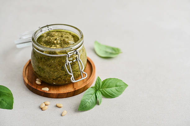 Sauce pesto in glass jar with fresh basil, hard cheese and pine nuts on light background