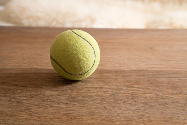 A vibrant green tennis ball with black seams sits on a textured wooden table, bathed in soft light.