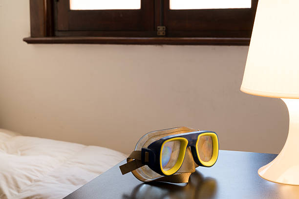 A classic yellow and blue scuba mask rests on a dark nightstand, illuminated by the warm glow of a table lamp. In the background, a cozy bed with a white duvet and a dark wooden window frame add to the relaxed, post-adventure atmosphere.