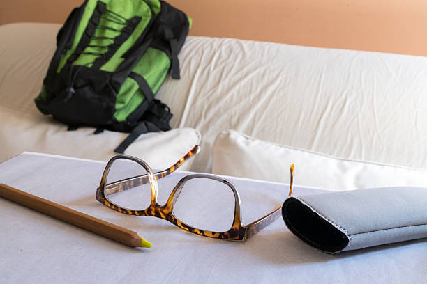 A pair of tortoiseshell glasses next to their case and a wooden pencil on a table, with a green backpack on a light-colored sofa in the background.