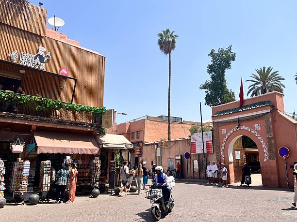 Marrakech, Morocco - June 5, 2025: People walking past street vendors and a traditional Moroccan archway on a sunny day, with a scooter driving by, capturing daily life in Rue Tougma, Marrakech