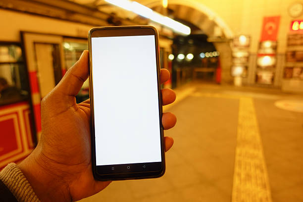 A person is holding a blank smartphone in a subway station during nighttime.