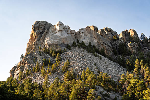 Aerial view of Mount Rushmore National Memorial surrounded by forest and granite cliffs under the warm light of sunset. Iconic landmark glowing in evening tones.