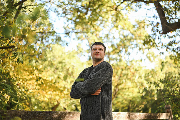 Man in a sweater standing outdoors with arms crossed, looking up with a confident expression