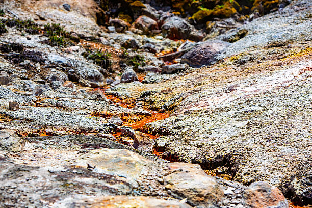A slender vein of rust-orange hydrothermal runoff snakes through pale, knobby sinter and scattered rocks in Yellowstone. Mineral deposits and heat-loving microbes tint the trickle vivid orange against the gray crust, with tiny bubbles and wet sparkle catching the sun.