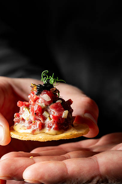Elegant close-up of a hand holding a cracker topped with gourmet steak tartare, garnished with black caviar and microgreens, on a dark background